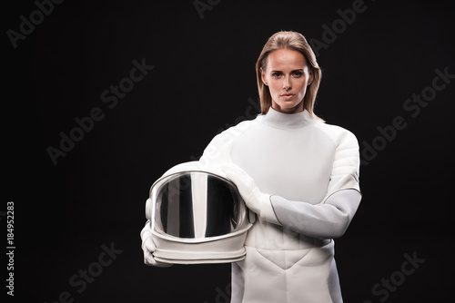 Cuadros en Lienzo Portrait of confident young female cosmonaut wearing hyperbaric astronaut protective suit is standing and looking at camera seriously while holding helmet