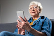 © Yakobchuk Olena - Portrait of old smiling lady looking at screen of mobile phone while sitting on couch