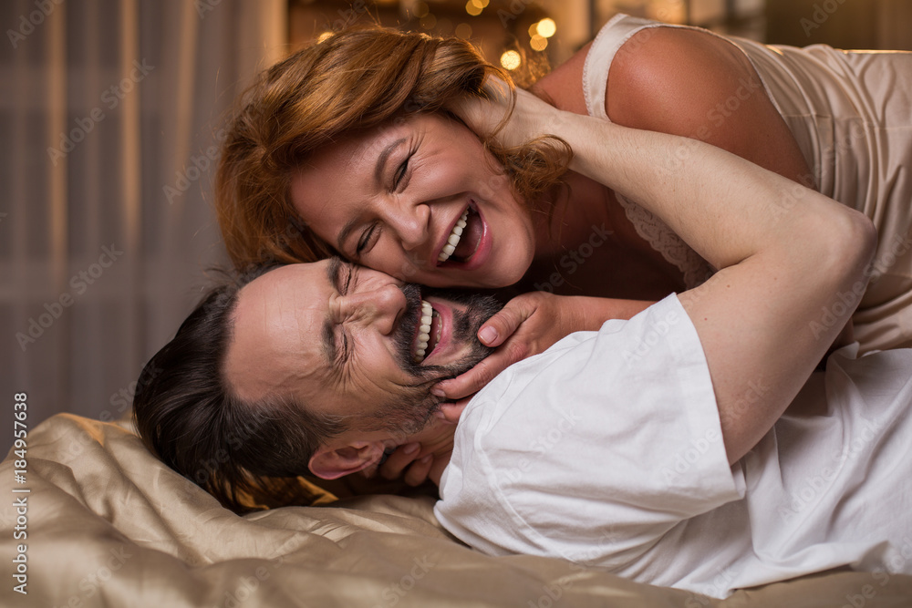Portrait of excited married couple lying on bed and laughing. They are hugging with closed eyes - obrazy, fototapety, plakaty 