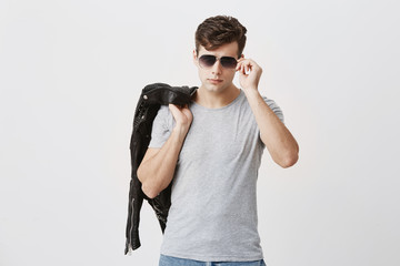 Wall Mural - Stylish, muscular, confident male posing against gray background. Attractive handsome caucasian guy with trendy haircut with black leather jacket thrown over shoulder, holding sunglasses in hand