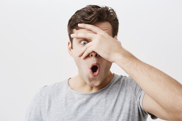 Wall Mural - Headshot of goofy surprised bug-eyed young european male student wearing casual grey t-shirt staring at camera with shocked look, expressing astonishment and shock, hiding face behind his palm.