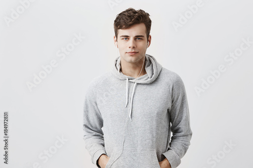 Fotografía Handsome attractive European man in gray hoodie, with hands in pockets, look pleased at camera, has good mood as comes home after work