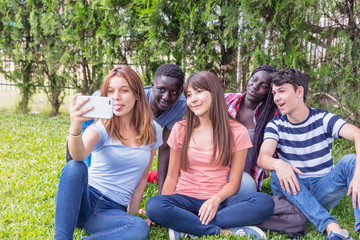 Sticker - Group of mixed races teenagers palying with smarthphone on the grass making selfies