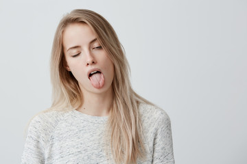 Wall Mural - Negative human reaction, feelings and attitude. Portrait of bored annoyed blonde girl in casual wear grimacing, sticking out her tongue, blinking, feeling nauseous because of bad smell or stink