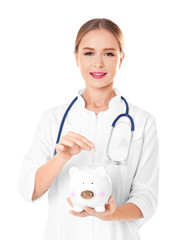 Canvas Print - Young female doctor putting coin into piggy bank on white background