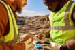 © Seventyfour - Mid-section portrait of two industrial workers wearing reflective jackets, one of them African, inspecting mineral ore on site outdoors and using digital tablet