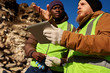 © Seventyfour - Low angle portrait of two industrial workers wearing reflective jackets, one of them African, inspecting mineral mines on worksite outdoors and using digital tablet