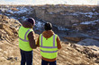 © Seventyfour - Back view portrait of two industrial workers wearing reflective jackets, one of them African, standing on cliff overlooking mineral mines on worksite outdoors, copy space
