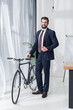 © LIGHTFIELD STUDIOS - smiling businessman in suit standing near bicycle in office