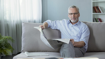 Senior man sitting on couch at home, holding newspaper and looking into camera