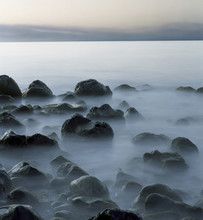 Tranquil View Of Rocks On Shore Against Sky During Foggy Weather