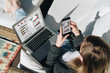 © foxyburrow - Sunny day.View from above.Young businesswoman is sitting on white couch,using laptop and smartphone with graphs, charts,diagrams on screen.Girl working,learning online.Distance work, online marketing.