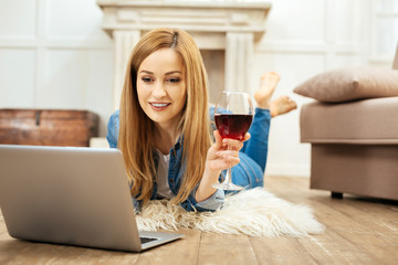 Great time. Beautiful dark-eyed blond smiling woman lying on the floor and holding a glass of red wine and working on her laptop