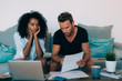 © DavidPrado - Young interracial couple in the couch stressed with financial problems doing calculations with paper work.