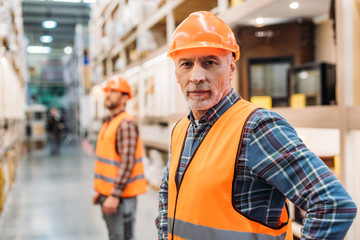 senior worker in safety vest and helmet, coworker standing behind in storage