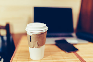 White paper Cup with burning coffee on the background of laptop and smartphone to Work on the go. Toned photo.