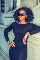 African American Woman casual street fashion. Wearing long sleeve dress, sunglasses, wristwatch, a black girl with braid hairstyle standing against vintage style wall, hand resting on hips, relaxing..