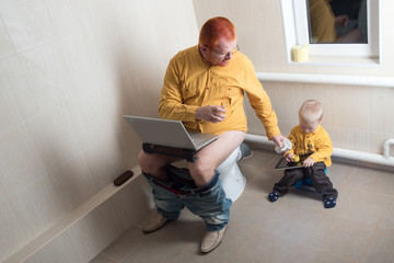 Man with red hair, beard and mustache in yellow shirt, glasses with laptop sitting on toilet bowl. Happy little kid on a potty