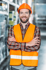 Wall Mural - cheerful worker in helmet and safety vest with crossed arms in warehouse