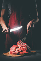 butcher with knife and fork cutting raw meat on wooden cutting board