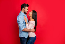Side View Of Caucasian, Attractive, Smiling Couple - Bearded Man Embrace His Charming, Cute, Pretty Woman, Looking To Each Other While Standing Over Red Background, 14 February