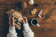Woman Holding Box With Christmas Cookies And Drinking Coffee On Wooden Table. Top View 