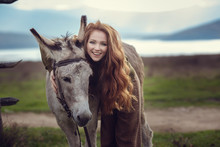 A Girl With Curly Red Hair In Fashionable Clothes In The Style Of Provence Hugs A Cute Donkey