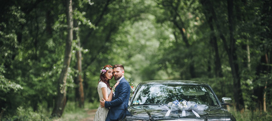 Wall Mural - Newlyweds near the black wedding car stand on the road in the summer forest. Panorama, copy space.