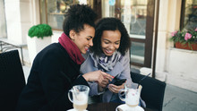 Two Attractive Mixed Race Female Friends Sharing Together Using Smartphone In Street Cafe Outdoors