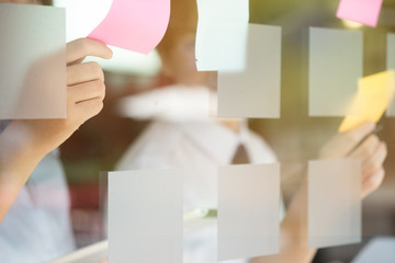 Creative business people reading sticky notes on glass wall with colleague working use post it notes to share idea discussing and teamwork, brainstorming concept.Closeup shot.