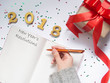 2018 New year's resolutions concept. Top view of women hands writing goal for new year or Christmas on opened blank notebook with gift box and party decorations on white table. Copy space. Flat lay.