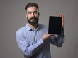 © sharplaninac - Happy smiling young bearded business person or programmer showing tablet screen with blank space for advertising against gray studio background.