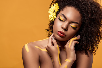 Young sensual african american woman with artistic make-up and gerbera in hair dreaming isolated on orange background