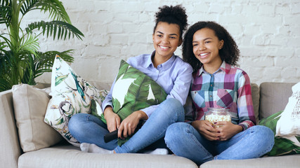 Wall Mural - Two mixed race curly girl friends sitting on the couch and watch comedy movie and eat popcorn. Funny sisters relaxing together at home