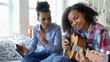 © silverkblack - Mixed race young woman with tablet computer sitting on bed teaching her teenage sister to play acoustic guitar at home