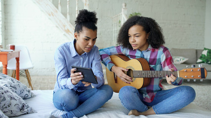 Mixed race young woman with tablet computer sitting on bed teaching her teenage sister to play acoustic guitar at home