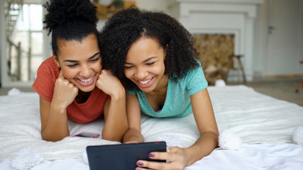 Wall Mural - Two cheerful mixed race funny girlfriends sharing social media using tablet computer and talks lying in bed at home