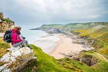 Female Backpacker Looking Into Distance At Rocky Sea Shore In Rhossili, Wales Coast Path