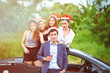 © Sasils Production - Christmas playboy party concept. Young white male stand on the side of convertible sport car holding pink champagne with four beautiful asian women in background wearing Santa hat.