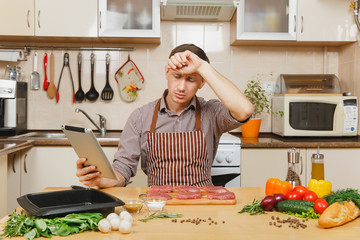 Tired European man in apron sitting at table with vegetables, looking for recipe in tablet, cooking at home preparing meat stake from pork, beef or lamb, in light kitchen full of fancy kitchenware.