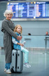 © Yakobchuk Olena - Full length portrait of happy gray-haired grandmother is hugging her little granddaughter from behind. They are looking at camera with joy while child is sitting on suitcase at airport. Copy space