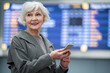 © Yakobchuk Olena - Portrait of joyful charming old woman is standing at international airport against electronic timetable and looking aside with happiness. She is keeping modern mobile phone. Copy space in right side