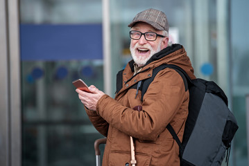 Fanny moments. Laughing old gray-haired man with beard is standing outdoors with backpack and mobile phone. He is looking at camera with wide smile