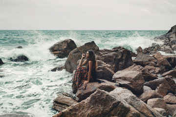 Wall Mural - Beautiful young boho styled woman sitting on a stone beach