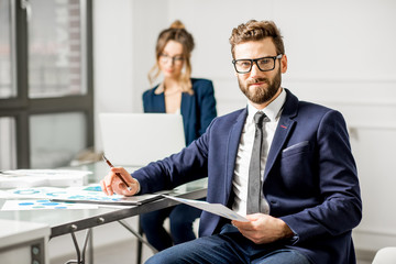 Portrait of a businessman dressed in suit working on documents with female assistant on the background at the white office interior