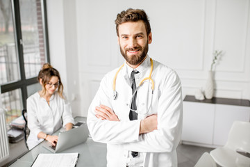 Portrait of an elegant senior doctor in medical gown with young female assistant on the background at the white office interior