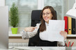© ViDi Studio - Beautiful smiling brown-hair business woman in suit and glasses sitting at the desk, working at computer with documents in light office, showing index finger on blank sheet. With place for text.