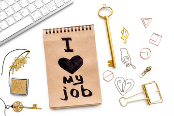 Happy at work. Handwritten motto I love my job in notebook near stationery on white background top view