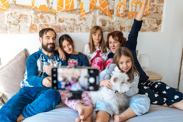 Wall Mural - Couple relaxed at home in bed with their three little daughters on the mobile phone taking a family selfie.