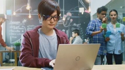 Talented Asian Developer Sits at the Table Works on a Laptop. In the Background Busy Office with Creative People Working on Personal Computers, Outlining Plan on a Glass Board with Sticky Papers. 